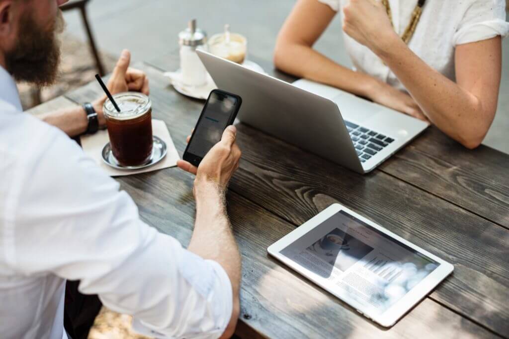 Male and female coworkers talking to one another across outdoor table with phone, tablet, and laptop in front of them