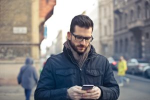 Bearded man in glasses and blue jacket checking SMS sent by business while crossing a city street