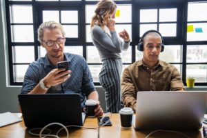 Business people at desk with woman behind them on phone.
