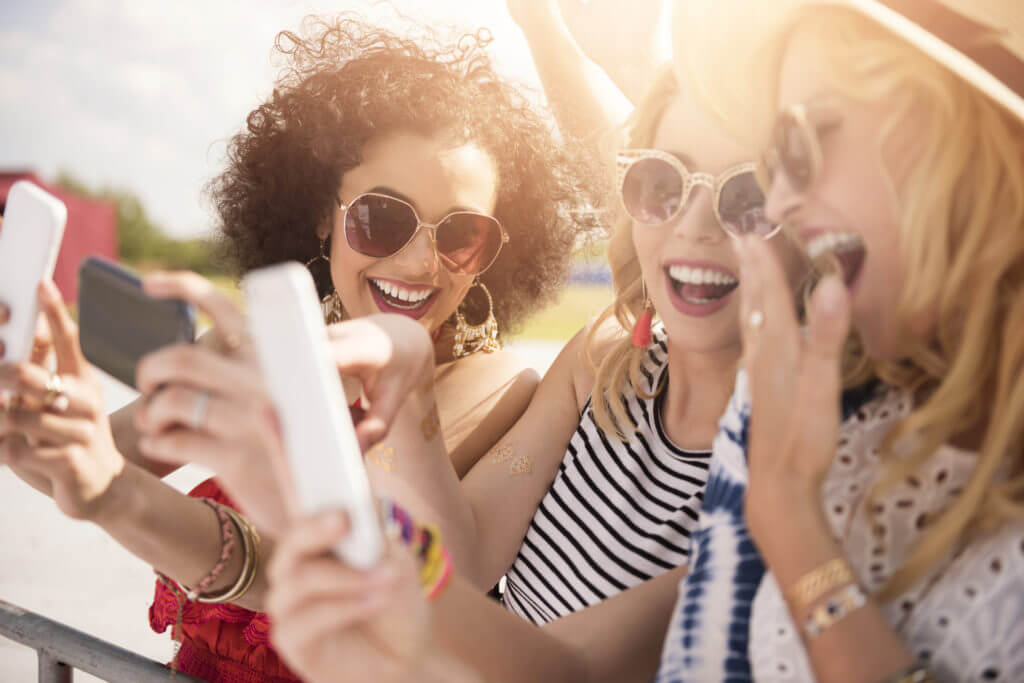 Three laughing women in sunglasses at an outdoor conference looking at their phones and grinning