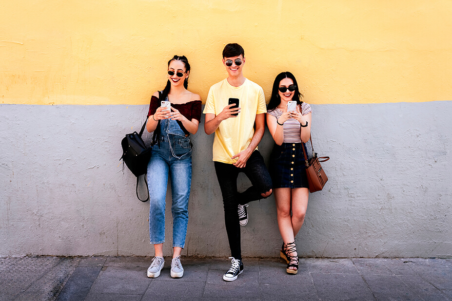 Three smiling shoppers leaning against a wall and reading retail texts on their phones